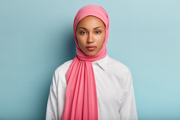 落ち着いた真面目な表情のアラビア人女性 肌の色が濃く ピンクのベールに包まれ 白いシャツを着て 化粧をしていない 自然の美しさ 青い壁のモデル イスラム教徒の女性のクローズアップショット 無料の写真