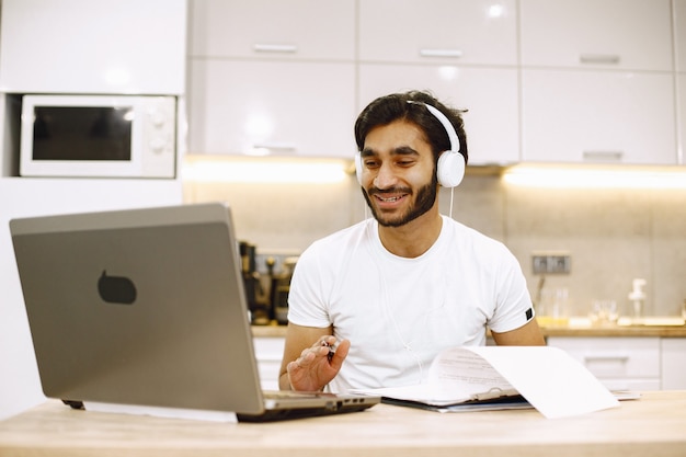 Arabic man watching online webinar, sitting in a kitchen with computer, enjoying distance learning. Free Photo