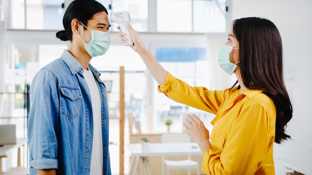 Asia female receptionist conducting wear protective face mask use infrared thermometer checker or temperature gun on customer's forehead before enter office. lifestyle new normal after corona virus. Free Photo