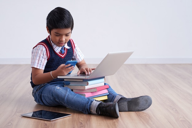Asian boy sitting on floor with tablet and laptop on stacked books, and using smartphone Free Photo