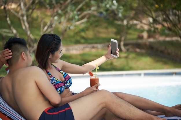 Asian couple lying on loungers by swimming pool and taking selfies on smartphone Free Photo