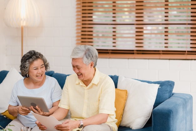 Asian elderly couple using tablet watching tv in living room at home, couple enjoy love moment while lying on sofa when relaxed at home. Free Photo