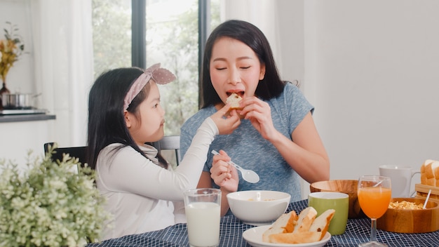 アジア系の日本人家族は家で朝食をとります アジアの母と娘が一緒に幸せな話をしながらパンを食べて オレンジジュース コーンフレークシリアル 牛乳をモダンなキッチンのテーブルの上の朝に飲みます 無料の写真