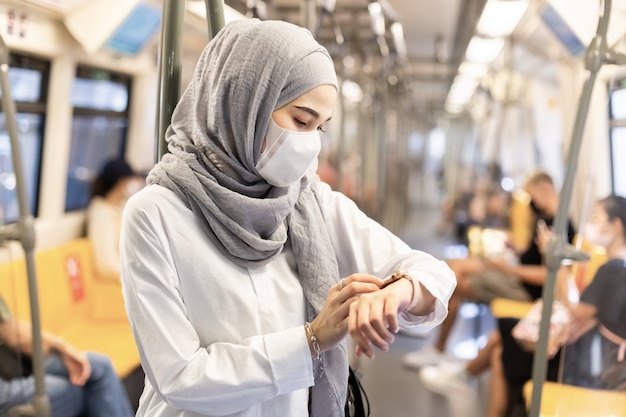 Asian muslim woman wearing medical face mask for prevent dust and infection virus and looking smartwatch in skytrain transit system public. Premium Photo