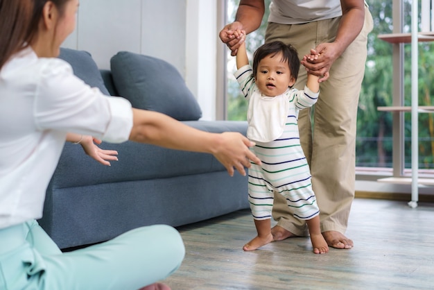 最初の一歩を踏み出すアジアの息子の赤ちゃんは彼の母親に向かって歩きます 父親の助けを借りて歩くことを学び 家で優しく歩く方法を教える幸せな小さな赤ちゃん プレミアム写真