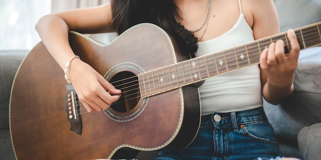 自宅でギターで音楽を演奏するアジアの女性 家の部屋で趣味で音を出す歌を演奏して歌うために座っている音響楽器を持った若い女性ギタリストミュージシャンのライフスタイル プレミアム写真