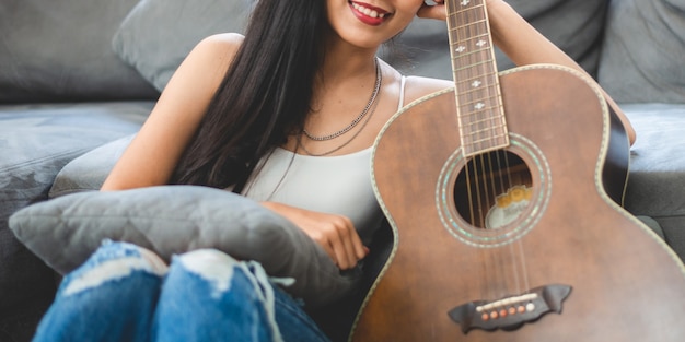 自宅でギターで音楽を演奏するアジアの女性 家の部屋で趣味で音を出す歌を演奏して歌うために座っている音響楽器を持った若い女性ギタリストミュージシャンのライフスタイル プレミアム写真