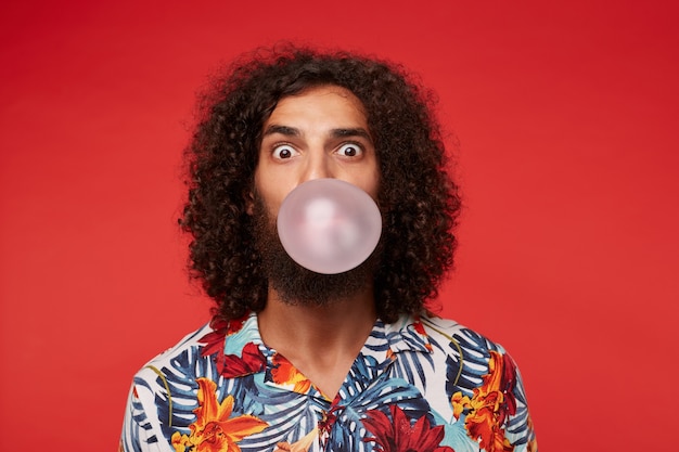 Astonished attractive young brunette curly man with beard wearing multi-colored flowered shirt while posing, rounding eyes and inflating bubble with pink gum Free Photo