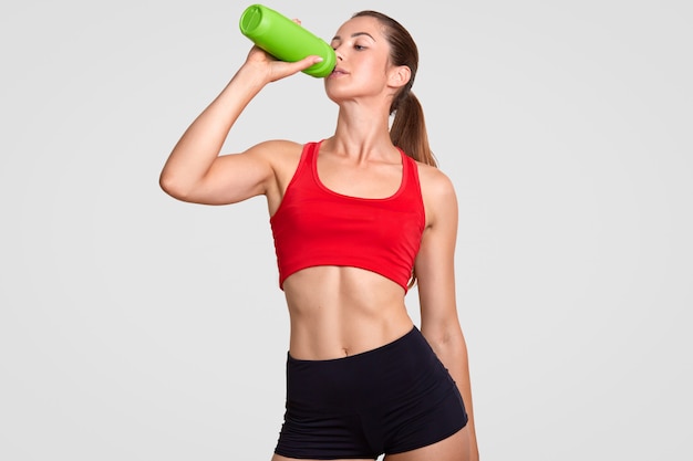 Athlete female drinks water, being thirsty after jogging during sunny day, has slim body, dressed in casual top and shorts, poses against white wall. people, energy and exhaustion concept Premium Photo