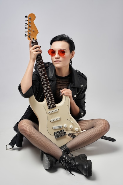白い背景の上のスタジオで革のジャケットと赤い眼鏡のエレキギターを持つ魅力的な女の子 プレミアム写真