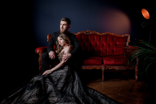 Attractive woman dressed in elegant evening dress and bearded handsome dressed in black suit holding hands together and sitting on the luxury red sofa Free Photo