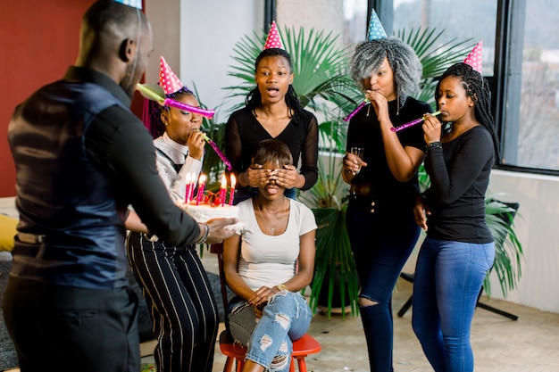 彼女の周りの彼女の友達と一緒に椅子に座って 彼女の誕生日の蝋燭を祝って 吹いて黒髪の魅力的な若いアフリカ女性 アフリカの男がキャンドルで誕生日ケーキを運ぶ プレミアム写真