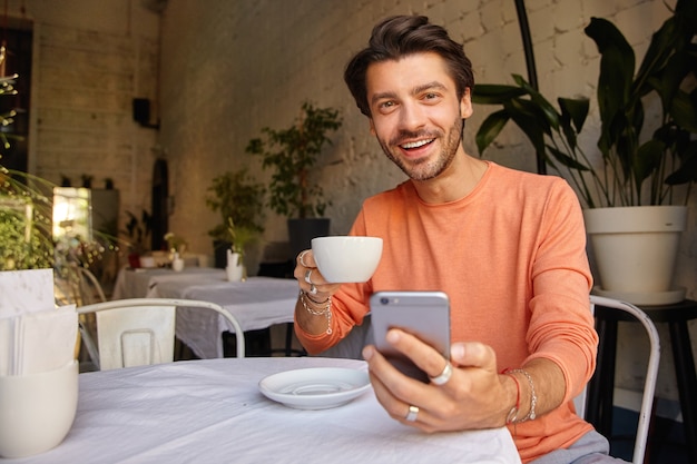 魅力的な若い黒髪の男性がカフェのインテリアにポーズをとって 手にsrartphoneを持って 元気に見て コーヒーを飲みながら 無料の写真