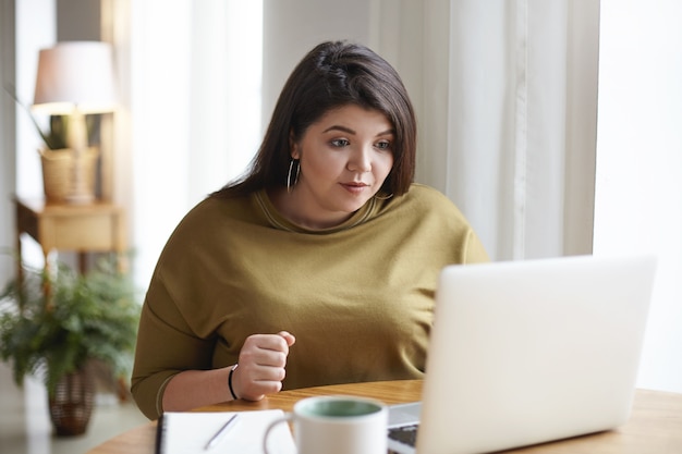 Attractive young dark haired woman with curvy body using generic laptop for remote work, drinking coffee, looking at screen with concentrated focused facial expression. technology and lifestyle Free Photo