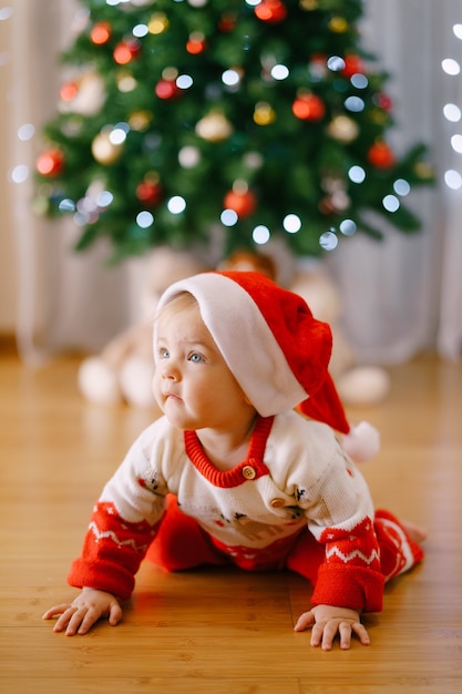 クリスマスの衣装を着た赤ちゃんとサンタの帽子がクリスマスツリーの前を這っています 高品質の写真 プレミアム写真