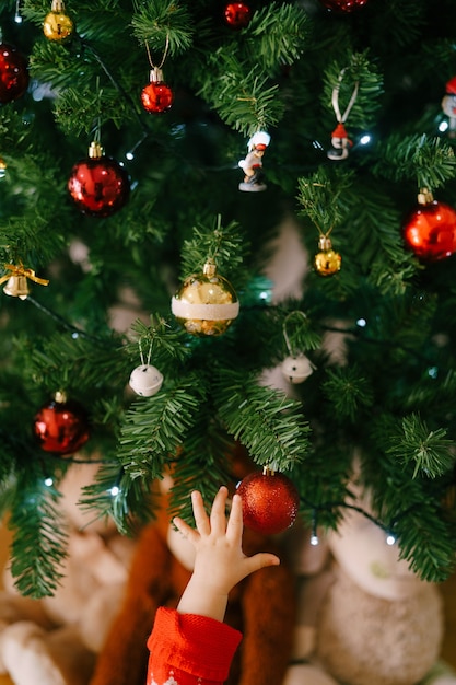 クリスマスツリーの赤いクリスマス飾りに手を伸ばす赤ちゃんの手 高品質の写真 プレミアム写真