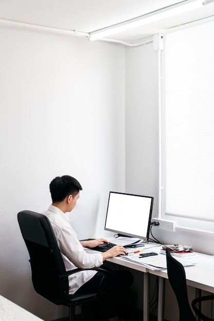 白いシャツを着て オフィスのテーブルに表示されている彼のパソコンでの作業 黒いオフィスの椅子に座る男の裏 プレミアム写真