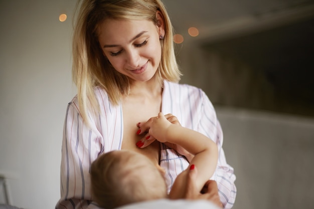 Back view of adorable six month old infant drinking breast milk. attractive young european woman in home clothing cradling her baby daughter in arms, breastfeeding her, enjoying deep connection Free Photo