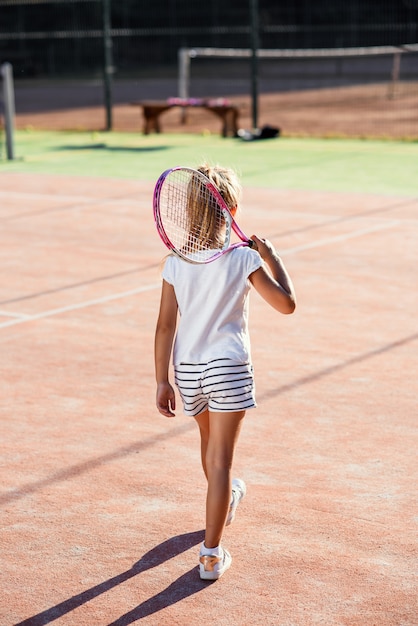 プレミアム写真 夕暮れ時の屋外テニスコートの上を歩いて肩にテニスラケットで白い制服を着た少女の背面図