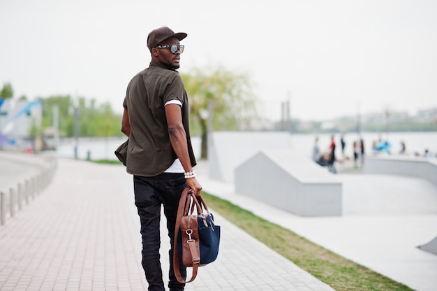 サングラスと屋外のハンドバッグとキャップにスタイリッシュなアフリカ系アメリカ人男性を着て歩くの背面図の肖像画 ストリートファッションの黒人男性 プレミアム写真