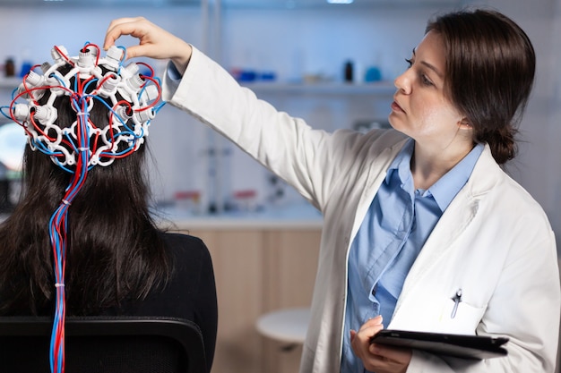 Back view of woman patient wearing performant eeg headset sitting on chair in neurological research laboratory while medical researcher adjusting it, examining nervous system typing on tablet. Free Photo