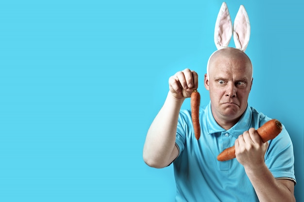 Bald brutal man in light t-shirt and rabbit ears. in his hands he holds the carrot of a different size. Premium Photo