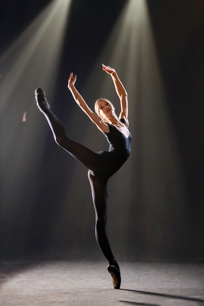 Photo | Ballerina in tight-fitting suit is dancing on black background on pointe shoes, is illuminated by sources of color.