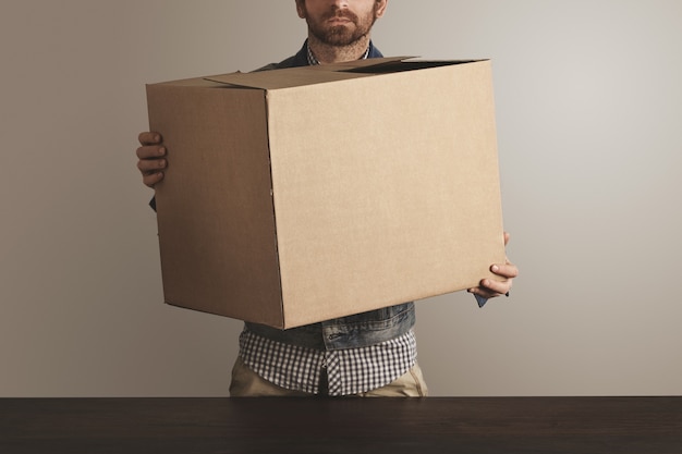 Bearded brutal courier in jeans work jacket holds big carton paper box with goods above wooden table. Free Photo