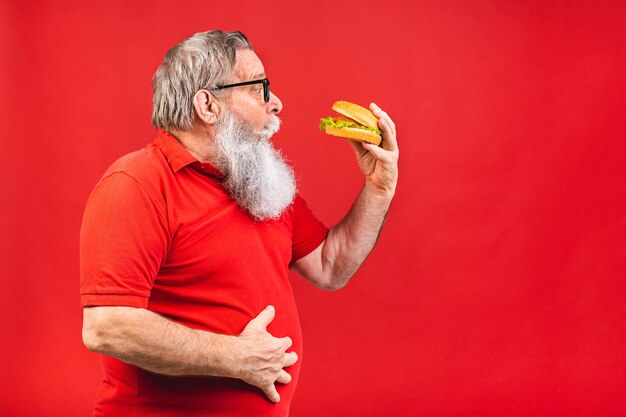 赤い背景で隔離のハンバーガーを食べる眼鏡とひげを生やした老人 プレミアム写真