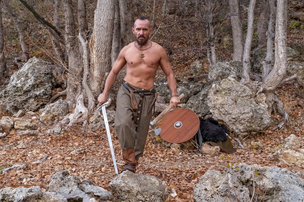 Premium Photo A Bearded Viking With A Naked Torso Holds A Sword In One Hand And A Battle Ax In