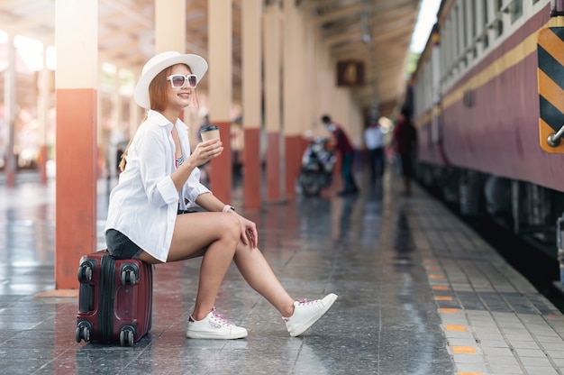 Beautiful asian traveler wearing hat and glasses holding cup of coffee while sitting on luggage at railway station, waiting for departure Premium Photo