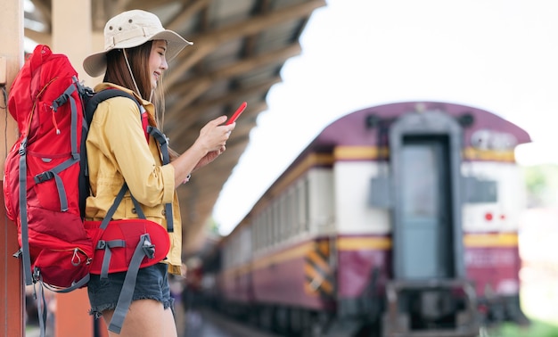 Beautiful asian traveler with backpack using smartphone while standing at railway station and waiting for departure Premium Photo