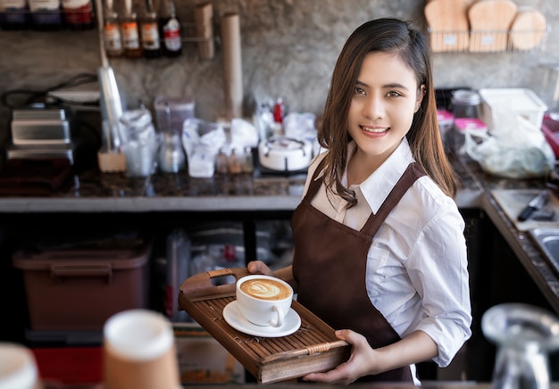 Premium Photo | Beautiful barista wearing brown apron holding hot coffee  cup served to customer with smili