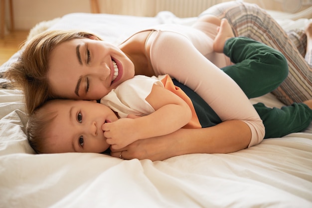 ゆったりとしたベッドに横になり 目覚めた幼児の息子を抱きしめて笑っている美しいブロンドの女性 寝室でかわいいお母さんと小さな子供の絆の居心地の良い甘いショット 家族 愛 ケア 愛情 無料の写真