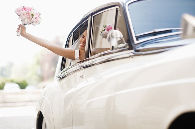 Beautiful bride sits with wedding bouquet in a retro car and has fun Free Photo