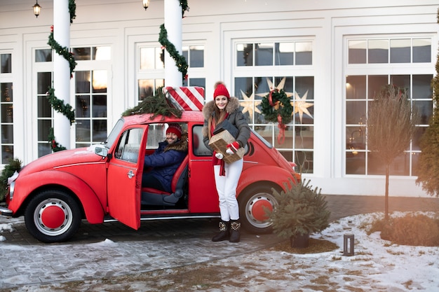 暖かい冬の服を着た美しい白人の女の子は彼女の夫に赤い車でクリスマスプレゼントの箱を運ぶ 無料の写真