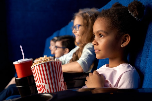 映画館で映画を見て面白い髪型で美しい集中アフリカの女の子 友達と一緒に座って ポップコーンを食べて 笑顔の愛らしい小さな女性の子供 プレミアム写真