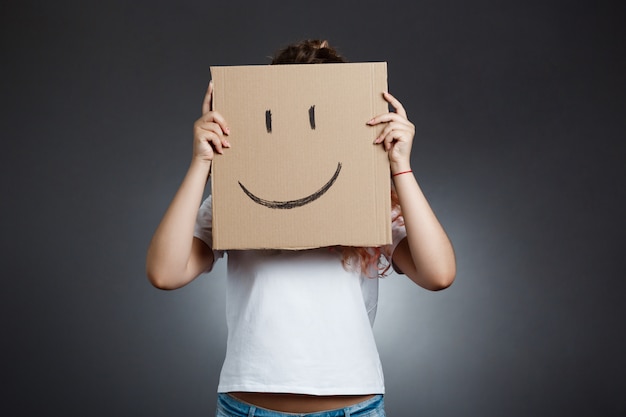 Beautiful girl hiding behind carton with smiley over grey wall. Free Photo