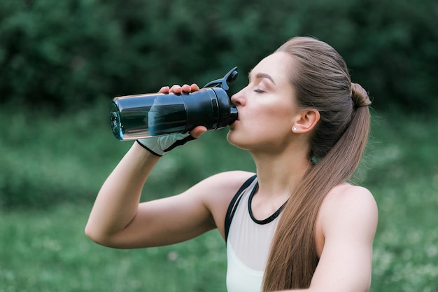 beautiful girl in sport clothes drinking water after workout while sitting on the grass Free Photo