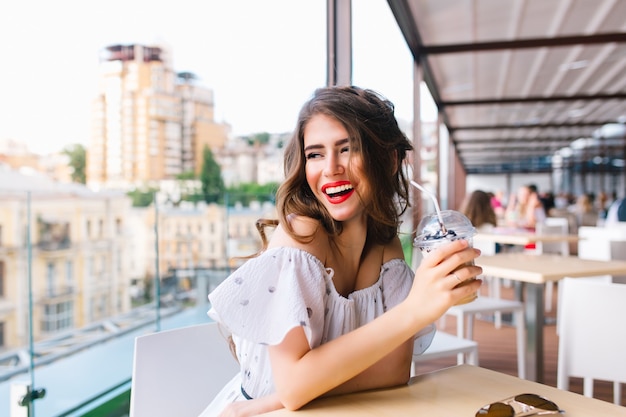 Beautiful girl with long hair is sitting at  table on the terrace in cafe . she wears a white dress with naked shoulders and red lipstick . she holds cup to go and smiles to the side. Free Photo