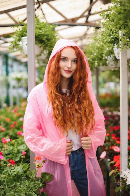 빨간 머리 곱슬머리를 한 아름다운 여인은 분홍색 비옷을 입고 온실에서 시간을 보내는 동안 카메라를 꿈꾸며 바라보고 있습니다 무료 사진