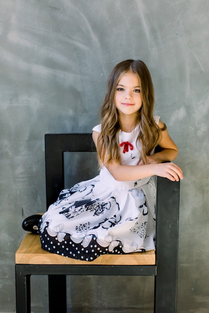 Premium Photo Beautiful Little Girl With Long Brown Hair And Blue Eyes Sitting On The Chair On A Grey Wall Wearing A Black And White Spring Dress