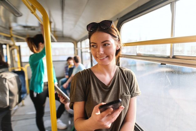 茶色の髪とサングラスの市バスに立って スマートフォンを使用して頭の上の美しい笑顔の白人少女 プレミアム写真