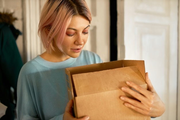Beautiful young european woman posing indoors with cardboard box in her hands, opening it, looking inside. Free Photo