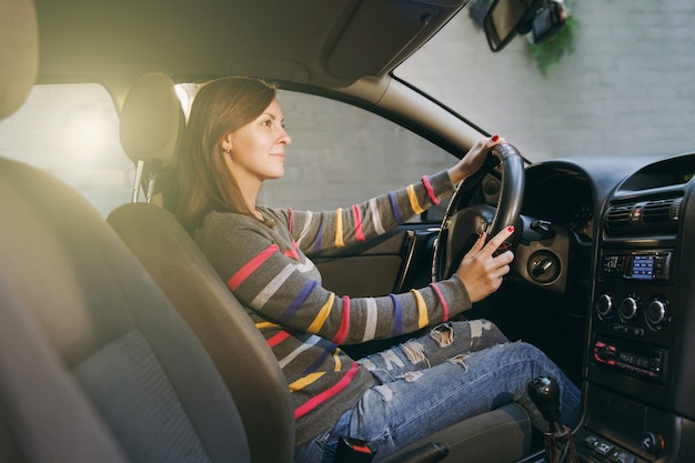 A beautiful young happy smiling european brown-haired woman with healthy clean skin dressed in a striped t-shirt sits in her car with black interior. traveling and driving concept. Free Photo