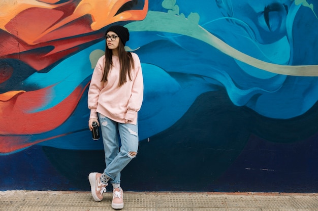Beautiful young woman holding aerosol can standing in front of colorful graffiti wall Free Photo