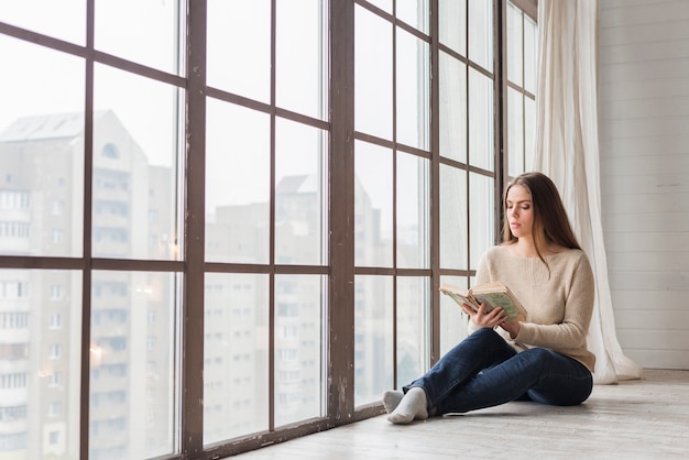 Beautiful young woman sitting near the glass window reading book | Free