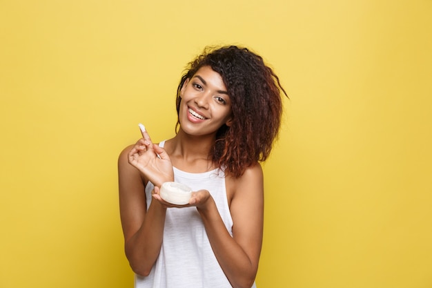 Beauty Concept - Beautiful young African American woman happy using skin care cream. Yellow studio background. Copy Space. Free Photo