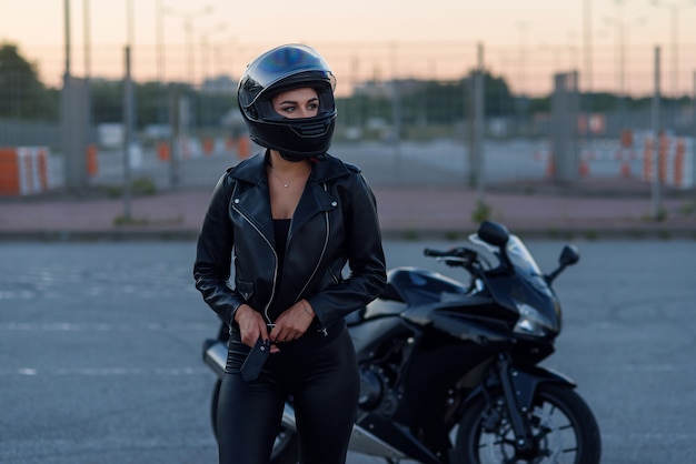 Premium Photo Biker Woman In Black Leather Jacket And Full Face Helmet Stands Near Stylish Sports Motorcycle Urban Parking Sunset In Big City