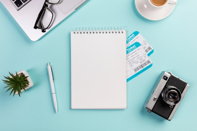 Blank spiral notepad with air tickets surrounded with laptop,eyeglasses,pen,camera,coffee cup on blue desk Free Photo